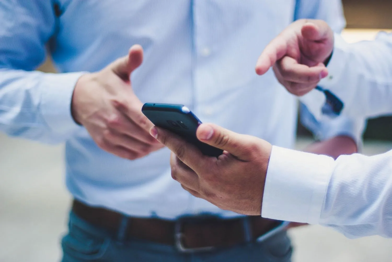 Smartphone, w dłoni mężczyzny w białej koszuli, z tyłu widać mężczyznę w niebieskiej koszuli.