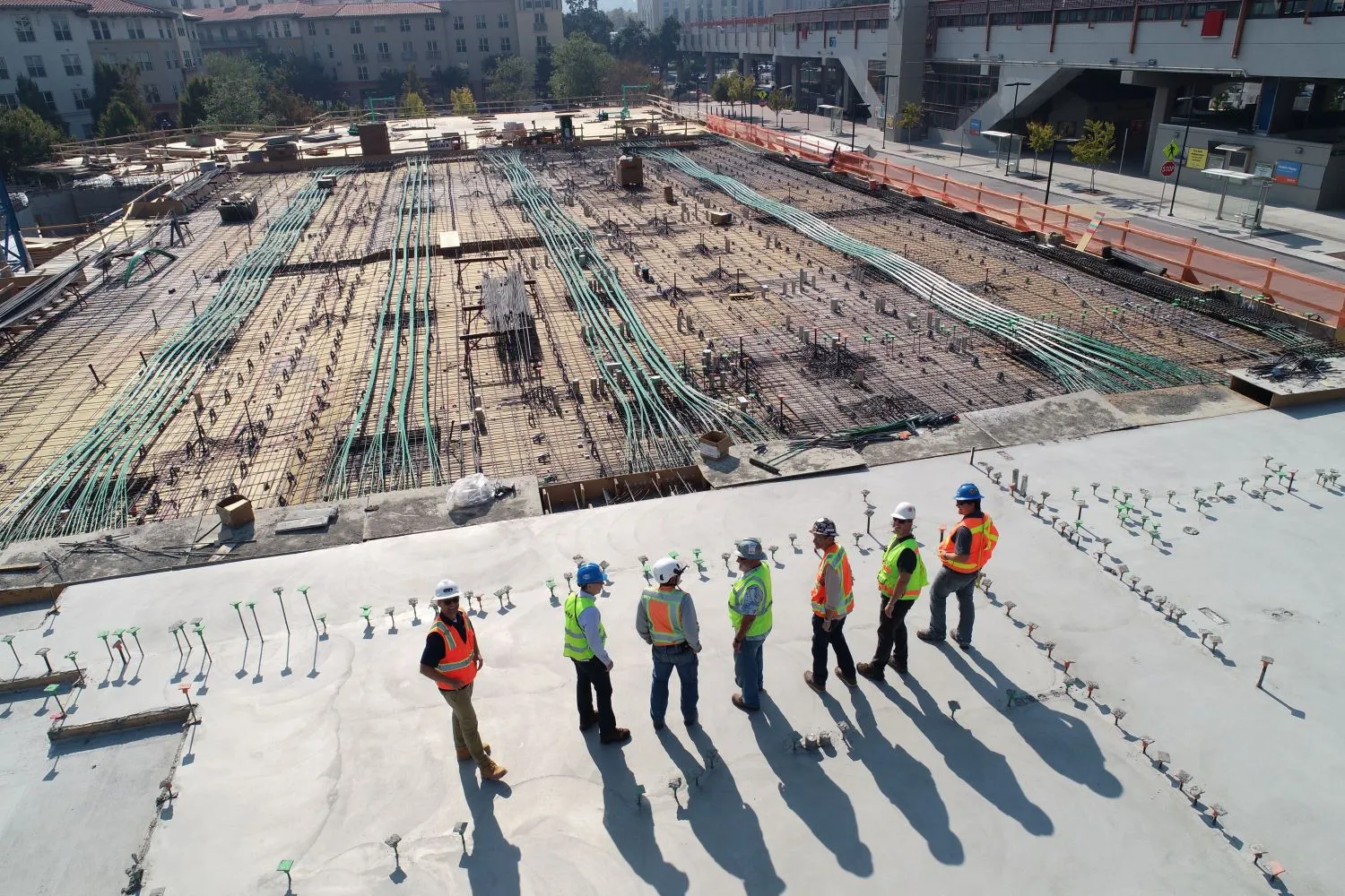 Siedmiu robotników w kaskach i kamizelkach odblaskowych, na betonowej wylewce. W tle, plac budowy, z widocznymi kablami.