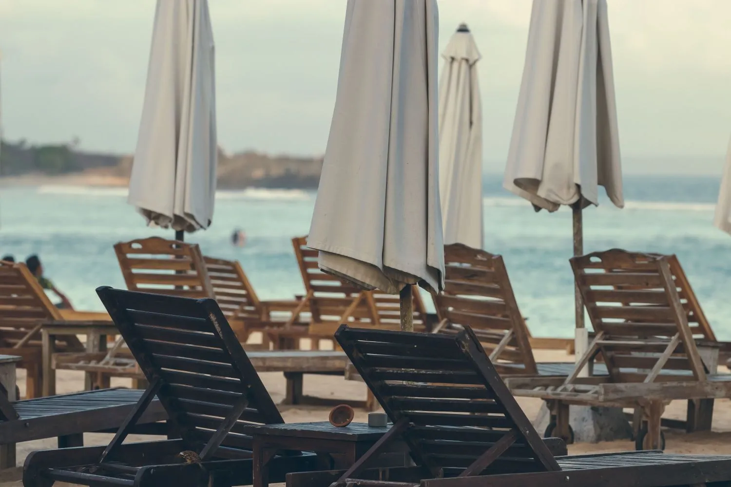 Drewniane leżaki oraz zwinięte parasole, stojące na plaży, w tle widać morze.
